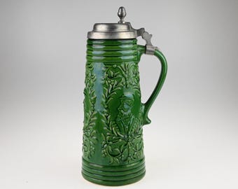 May include: A tall, cylindrical green ceramic beer stein featuring a relief-molded design of a bearded figure surrounded by intricate floral vines. The vessel includes a metallic pewter lid with a thumb-operated hinge and a matching green handle.