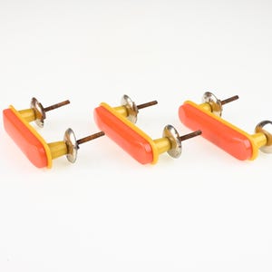 Könnte beinhalten: Drei Schubladengriffe mit einer rechteckigen orangefarbenen Vorderseite sowie gelben Seiten und Unterseiten. Jeder Griff verfügt über zwei silberfarbene Metallbefestigungen mit Gewindeschrauben an der Rückseite zur Montage an Möbeln.