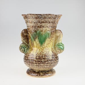May include: A decorative ceramic vase with a textured brown, tan, and green glaze. The vase has two ornate handles and a ruffled rim. The base is round and the vase is approximately 8 inches tall.