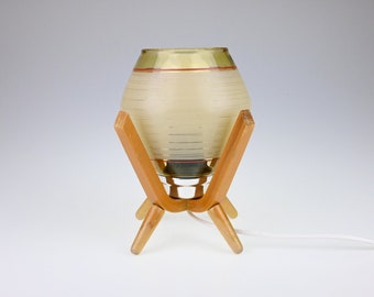 Puede incluir: Una lámpara de mesa moderna de mediados de siglo con una pantalla de vidrio blanco y una base de madera. La pantalla tiene una franja dorada y roja alrededor de la parte superior.