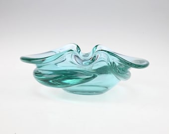 May include: A translucent light teal glass bowl featuring an organic, free-form shape with undulating, wavy edges. The glass has a smooth, polished surface and a thick, rounded rim that curves inward and outward to create a fluid silhouette.