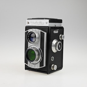 May include: A black twin-lens reflex camera with silver metal accents and the brand name Wellaflex printed on the top front panel. The camera features two circular lenses, a side-mounted focus knob, and various mechanical dials and settings.