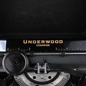 May include: Close-up of a vintage black Underwood Champion typewriter. The name "UNDERWOOD CHAMPION" is printed in gold lettering on a black bar. The typewriter's keys, rollers, and other mechanical components are visible, showcasing its intricate design.