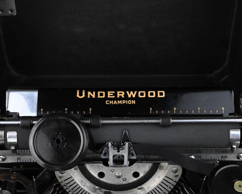May include: Close-up of a vintage black Underwood Champion typewriter. The name "UNDERWOOD CHAMPION" is printed in gold lettering on a black bar. The typewriter's keys, rollers, and other mechanical components are visible, showcasing its intricate design.
