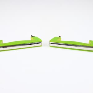 May include: Two lime green drawer handles with a horizontal silver-toned metal strip running along the front face. Each handle features two small black mounting posts extending from the back, designed for attachment to a flat surface or furniture.