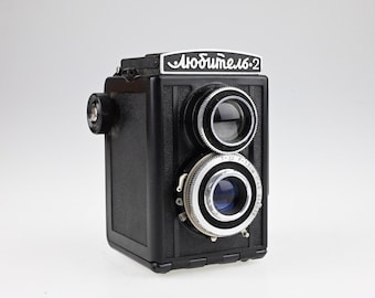 May include: A black twin-lens reflex camera featuring a top viewfinder lens and a bottom taking lens with silver metal rings. The top plate displays the Cyrillic text "Любитель-2" in white on a black background, while the lens barrel shows technical markings.