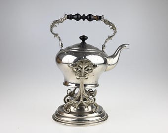 May include: A silver-plated tea kettle with a polished finish, featuring a black wooden handle and a matching black knob on the lid. The kettle rests on an ornate, decorative silver-plated metal stand with a curved spout and intricate detailing.
