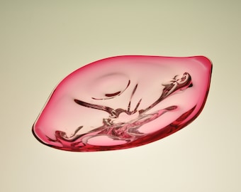 May include: A pink glass bowl with a unique, organic shape. The bowl is made of clear glass with a pink tint and has a wavy, flowing design.
