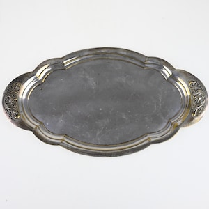 Può includere: Un vassoio da portata ovale in metallo color argento con bordo smerlato e una superficie centrale liscia e riflettente. Le due maniglie presentano un motivo floreale in rilievo con sottili accenti dorati, creando un contrasto decorativo.