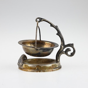 Puede incluir: Un infusor de té plateado con una cesta colgante y un soporte decorativo. El soporte tiene un diseño floral y un asa curva.
