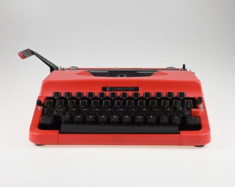 May include: A bright red Chevron typewriter featuring a black keyboard with white lettering, a black carriage return lever with a red tip, and a prominent black space bar. The brand name Chevron is printed in white on the front center panel.