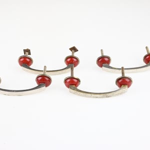 Op de afbeelding: Drie zilverkleurige metalen ladehandgrepen met een gebogen, boogvormig ontwerp. Elke handgreep is voorzien van twee felrode, ronde decoratieve accenten aan de uiteinden en bevat twee metalen schroeven met schroefdraad voor bevestiging aan meubels.