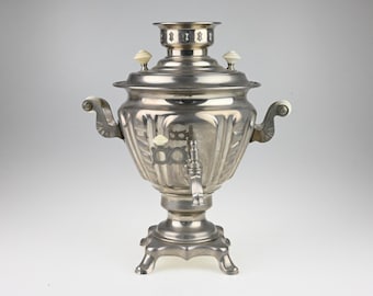 May include: A metallic silver samovar featuring a fluted body, a central spigot with a lever, and two side handles. The vessel includes a top lid with small white knobs, a wide base with four feet, and a decorative collar at the neck.