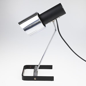 Könnte beinhalten: Eine schwarze und verchromte Schreibtischlampe mit zylindrischem Schirm und einem gebogenen, silberfarbenen Stützarm. Die Lampe hat einen rechteckigen Sockel und ein schwarzes Netzkabel. Die Lampe ist für den Schreibtischgebrauch konzipiert.