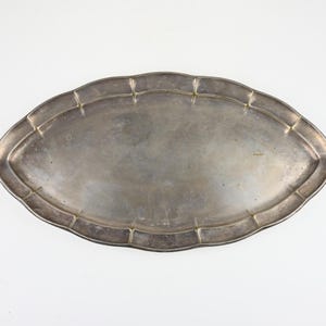 Puede incluir: Una bandeja de servir ovalada de color plateado con un borde festoneado. La bandeja tiene un diseño simple y elegante, con un borde ligeramente elevado y detalles decorativos a lo largo del borde. La superficie de la bandeja tiene un acabado pulido.
