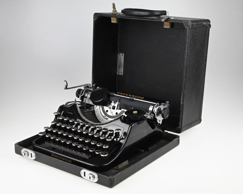 May include: A vintage black Underwood typewriter with silver keys and accents, displayed in its open black carrying case. The typewriter features a round key design and the brand name is visible on the front.