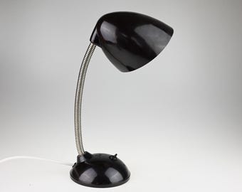 May include: A black desk lamp featuring a glossy, dome-shaped shade and a flexible, silver-toned metal gooseneck arm. The lamp is supported by a round, black base that includes a small toggle switch and a white power cord extending from the back.