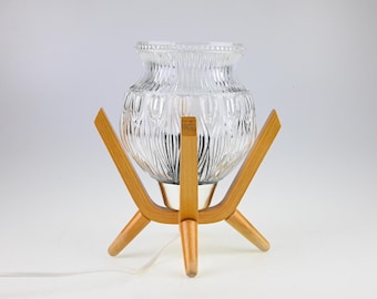 May include: A table lamp featuring a clear, textured glass shade with a vertical ribbed pattern, supported by a light-toned wooden tripod base with three angled legs. The lamp includes a white electrical cord extending from the base.