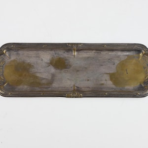May include: An elongated rectangular metal tray with rounded corners and a tarnished brass finish. The tray features decorative embossed floral motifs at both ends and along the edges, with a smooth, worn central surface showing dark patina.