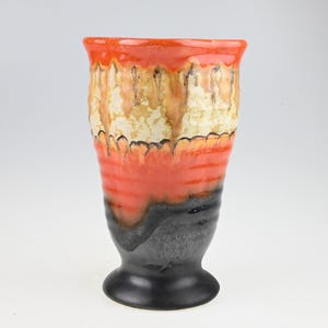 May include: A tall, ceramic vase with a flared rim and a pedestal base. The vase features a layered design with colors including orange, cream, and black. The exterior has a textured, glazed finish, adding visual interest to the piece.