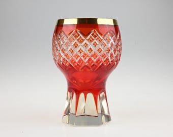 May include: A red and clear cut crystal glass featuring a diamond-patterned upper section and a smooth, faceted base. The rim of the glass is finished with a thin, polished gold band, providing a distinct contrast to the vibrant red glass body.