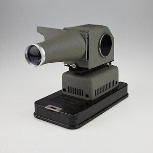May include: Vintage gray slide projector with a black base. The projector has a lens with a silver rim and a cooling vent on the side. The device is designed to project images from slides onto a screen.