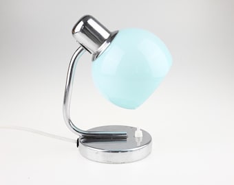 May include: A table lamp featuring a glossy, light blue, spherical glass shade attached to a polished chrome metal arm and a circular chrome base. A small white push-button switch is located on the top surface of the base, with a white power cord.