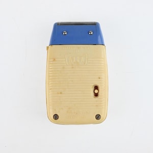 Puede incluir: Afeitadora eléctrica vintage con cuerpo color crema y parte superior azul. La afeitadora tiene forma rectangular con textura acanalada en la parte delantera. La marca "ALFA" es visible. Dos tornillos aseguran la base.
