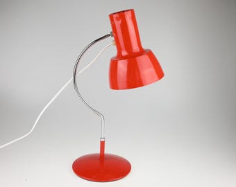 May include: A bright red desk lamp featuring a cylindrical head and a wide, flared shade. The lamp is supported by a curved, polished chrome arm that connects to a circular red base. A white power cord extends from the back of the lamp head.