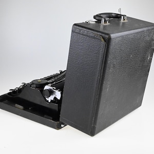 May include: A black vintage typewriter with a matching carrying case. The typewriter is open, revealing the keys and mechanical components. The case is upright, with a handle on top. The typewriter is on a white background.