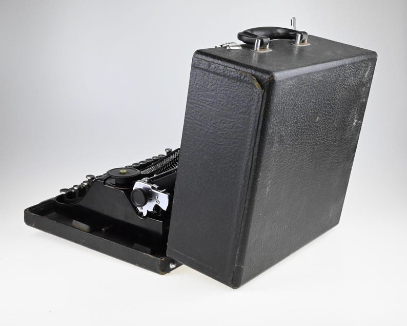 May include: A black vintage typewriter with a matching carrying case. The typewriter is open, revealing the keys and mechanical components. The case is upright, with a handle on top. The typewriter is on a white background.