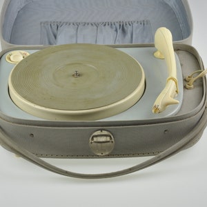 Op de afbeelding: Een vintage draagbare platenspeler in een grijze draagtas met een crèmekleurige draaitafel en toonarm. De tas heeft een handvat en een metalen sluiting.