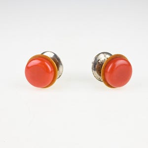 May include: A pair of round stud earrings featuring a smooth, vibrant orange cabochon centre surrounded by a thin mustard yellow border, set against a metallic silver-toned backing with a visible post mechanism for secure attachment.