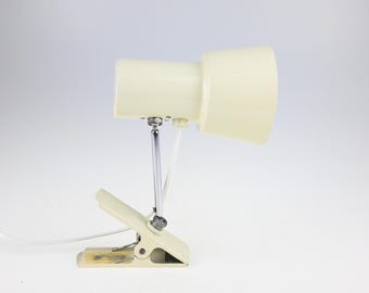 May include: A cream-colored clip-on desk lamp featuring a cylindrical head, a chrome-finished adjustable metal arm, and a sturdy spring-loaded base clamp. A white power cord extends from the back of the lamp head, providing a functional lighting solution.