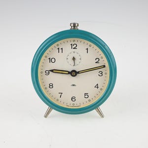 May include: A round analog alarm clock with a teal metal casing and a cream-colored face. The clock features black Arabic numerals, a small sub-dial at the top, and black hands with yellow centers. It stands on two small silver-toned metal legs.