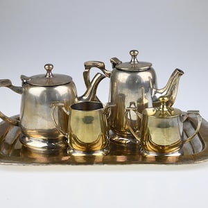 以下が含まれることがあります： トレイ付きの銀色のティーセット。セットには、ティーポット、クリーマー、砂糖入れ、サービングトレイが含まれています。ティーポットには蓋と曲がった注ぎ口が付いています。クリーマーと砂糖入れは金色です。トレイは長方形です。