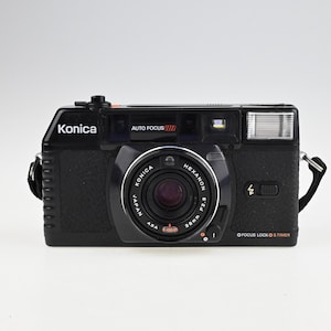 May include: A black Konica Auto Focus camera with a silver lens. The camera has a built-in flash and a focus lock button.