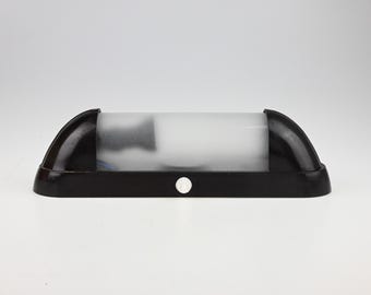 May include: A horizontal wall-mounted light fixture featuring a black base and a semi-cylindrical frosted glass shade. The design includes a small circular white switch centered on the front of the black frame, providing a clean and functional aesthetic.
