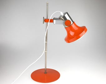 May include: A desk lamp featuring a bright orange circular base and a matching orange adjustable shade. The lamp has a vertical silver metal pole, a silver metal arm, and a white power cord. The shade is connected to the arm with orange circular hinges.
