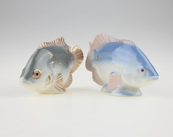 May include: Two ceramic fish figurines. The left fish is grey with gold-painted scale details and a pink eye. The right fish is light blue with a pinkish-tinted fin and tail. Both figurines have a smooth, glossy finish and stand on flat bases.