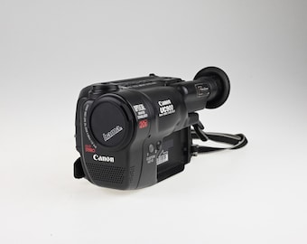 May include: A black Canon UC900 8mm video camcorder featuring a Hama lens cap, a 20x optical zoom lens, Hi-Fi stereo audio, and an optical image stabilizer. The device includes a viewfinder, custom keys, and an attached black carrying strap.