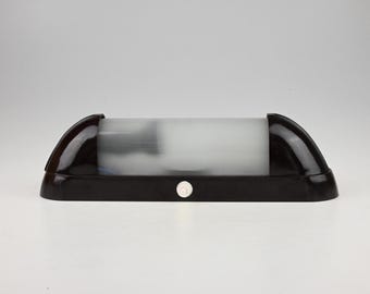 May include: A horizontal wall lamp featuring a cylindrical frosted glass shade held by two dark brown, curved end caps. The base is a matching dark brown rectangular frame with a small, circular white push-button switch centered on the front edge.