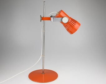May include: A retro-style orange desk lamp with a cylindrical head and a round base. The lamp features a silver metal arm and a white power cord. The lamp's head is angled downward, and the base is approximately 8 inches in diameter.