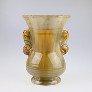 May include: A ceramic vase with a flared rim and two ornate handles. The vase has a glossy finish with shades of yellow, brown, and green. The base is round and the handles are detailed with floral accents. A decorative piece.