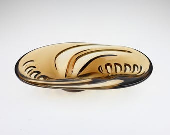 May include: An oval-shaped amber glass bowl featuring a smooth, translucent finish and a series of curved, recessed indentations along the interior rim that create a rhythmic, wave-like pattern across the surface of the glass vessel.