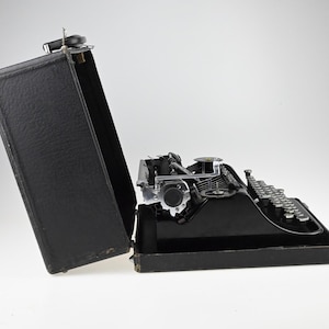 May include: A black vintage typewriter with silver keys and mechanical components, partially open in its black carrying case. The typewriter is set against a white background, showcasing its classic design.