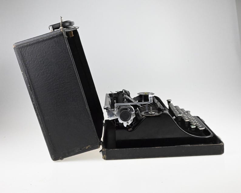 May include: A black vintage typewriter with silver keys and mechanical components, partially open in its black carrying case. The typewriter is set against a white background, showcasing its classic design.