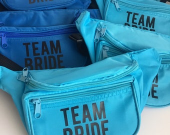 May include: A group of blue fanny packs with black zipper pulls and the text "TEAM BRIDE" printed on the front.