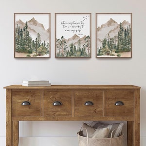 May include: Three framed watercolor prints depicting mountain and forest scenes. The center print features the quote "between every the pine trees, There is a door leading to a new way of life" by John Muir. The prints are displayed above a wooden console table.