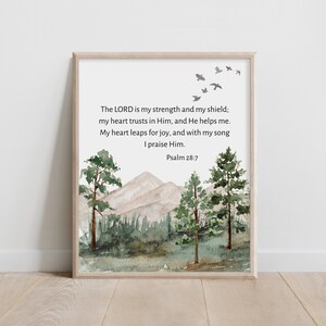 May include: A framed watercolor print featuring a serene landscape with mountains, trees, and flying birds. The artwork includes the text of Psalm 28:7, a verse from the Bible, in a simple, elegant font. The frame is a light wood color.